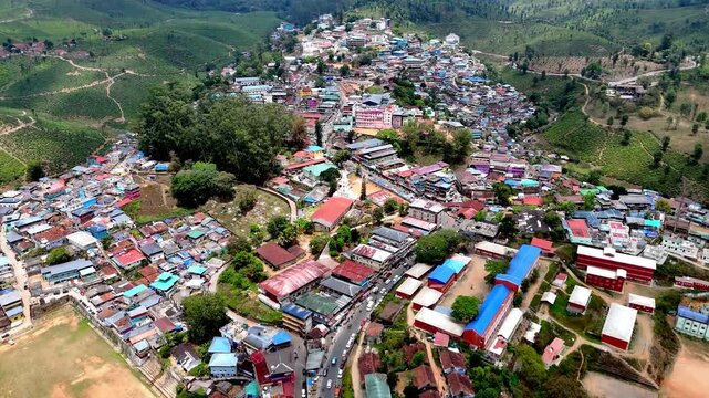 Smooth forward aerial view traveling above a compact town toward undulating hills, showcasing bright rooftops, open grounds, and surrounding greenery under dramatic atmospheric light.