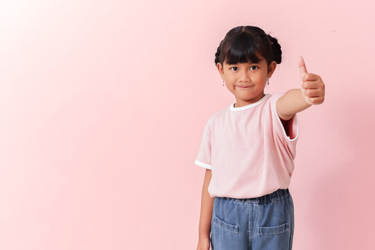 smiling young southeast asian girl in pink shirt giving thumbs up to camera on isolated pink background, cheerful approval gesture in clean studio portrait