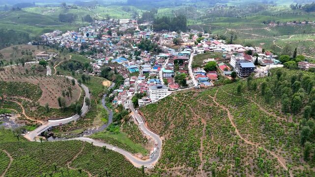 Aerial view of a vibrant hill town in summer, surrounded by lush tea estates and winding mountain roads. Colorful rooftops cluster along the slopes while a stream flows through the valley