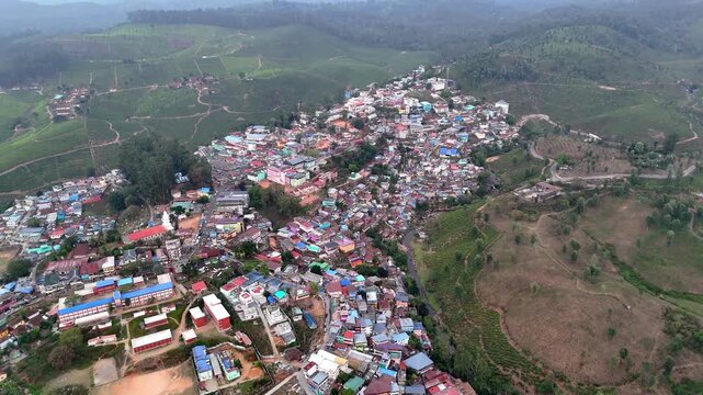 Drone shot capturing a compact settlement spread across uneven terrain, featuring vibrant houses, narrow streets, and surrounding hills with sparse vegetation and plantation patches.