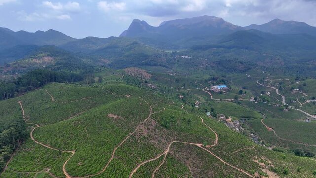 Backward-moving aerial footage revealing expansive green hills with winding dirt roads, gradually pulling away to unveil layered mountain ranges and a vast, scenic landscape under dramatic clouds.