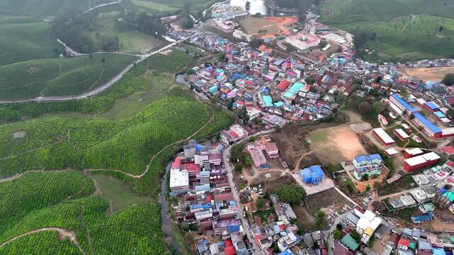Drone shot capturing a vibrant settlement with mixed residential and institutional buildings, central open spaces, winding streets, and surrounding greenery blending into the hilly landscape.