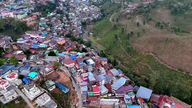 Drone footage moving forward across a vibrant settlement, transitioning from dense buildings to expansive green hills, with layered terrain and soft haze adding depth and motion.
