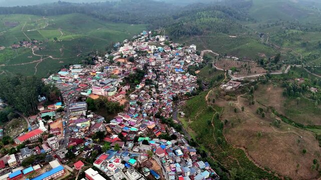 Cinematic aerial view of a dense hillside town with colorful rooftops clustered tightly together, surrounded by rolling green hills, winding roads, and patches of open land.