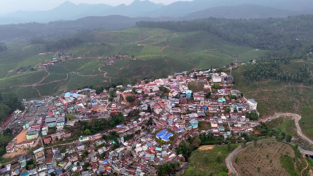 Wide cinematic aerial of a compact town highlighting varied architecture, bright rooftops, open fields, and road networks, bordered by lush plantations and undulating hills.
