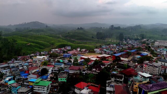 Cinematic forward-moving aerial shot gliding over a hillside town with colorful rooftops, gradually revealing lush green plantations and rolling hills ahead under a moody, cloud-filled sky.