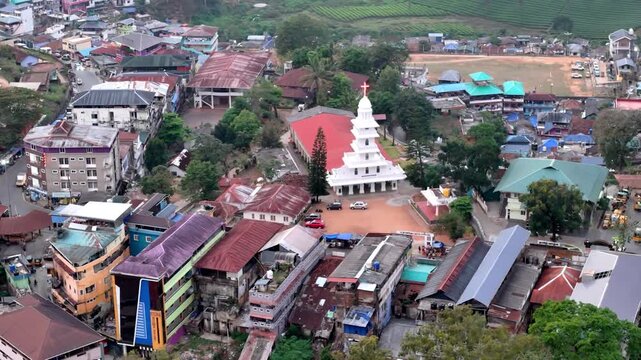 Cinematic aerial view of a hillside town centered around a striking white church with a red roof, surrounded by tightly packed buildings, narrow streets, and scattered greenery creating a peaceful com