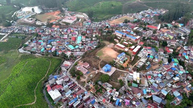 Cinematic aerial view of a hillside town showcasing colorful rooftops, organized clusters of buildings, open grounds, and curving roads, surrounded by green plantation areas and rolling terrain.