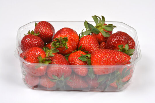 A transparent plastic punnet filled with ripe red strawberries with green leaves against a plain white background
