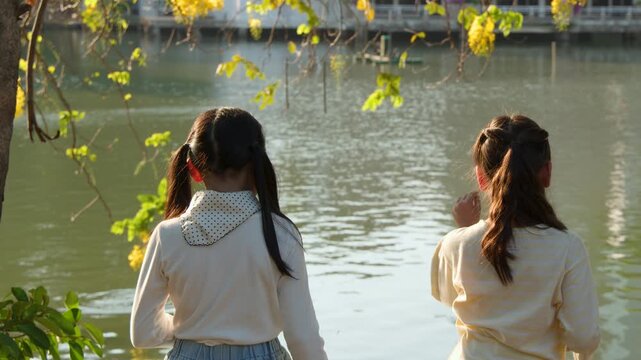 Two Young Thai Girls Feeding Fish in a Scenic Bangkok Park Pond