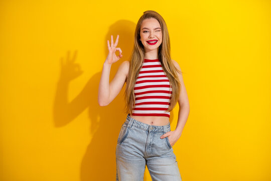 Cheerful young woman posing against yellow background with a playful OK gesture symbolizing positivity and style