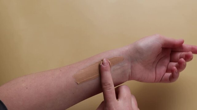 Woman applying liquid foundation on fingers, makeup routine with cosmetic concealer on beige background