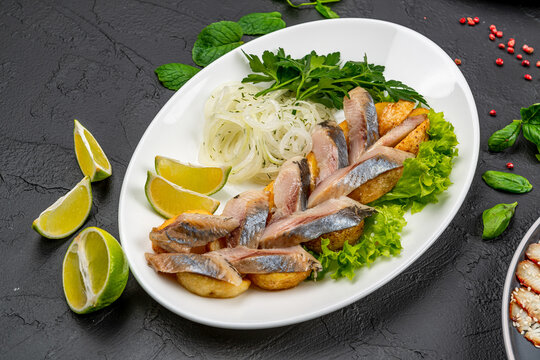 Sliced salted herring served on roasted potatoes with lime. White platter with marinated herring slices, fried potato wedges, onion rings, lime wedges, and fresh herbs on black.