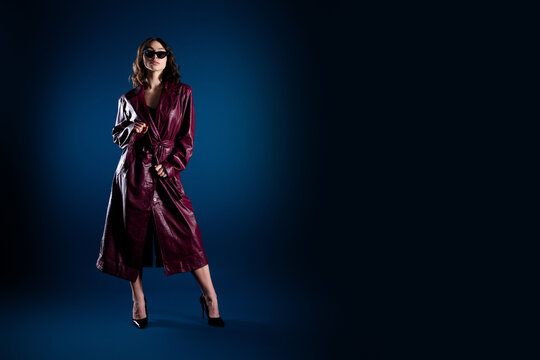 Fashion model in burgundy leather trench coat and sunglasses poses confidently with high heels in dramatic studio lighting