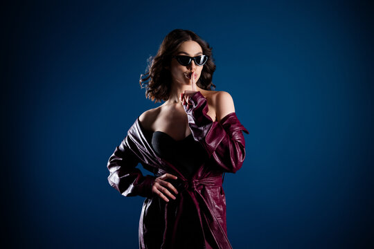 Fashionable woman in sunglasses and burgundy leather coat gestures for silence with finger to lips in moody studio portrait