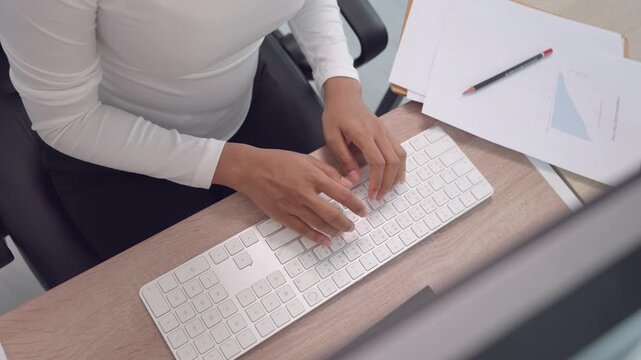 Top view of woman typing on keyboard with business report documents on office desk. Concept of data analysis, reporting, office productivity, and professional workflow management.