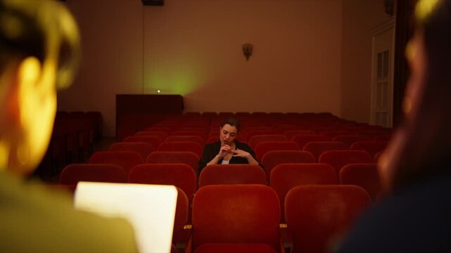 Solitary spectator in empty theater auditorium listening to presenters on stage, illuminated by warm cinematic lighting and surrounded by rows of red velvet seats