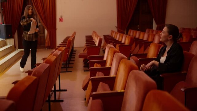 Young woman rehearsing alone in an empty theater auditorium, passionately gesturing and practicing lines between rows of red seats before a dramatic performance
