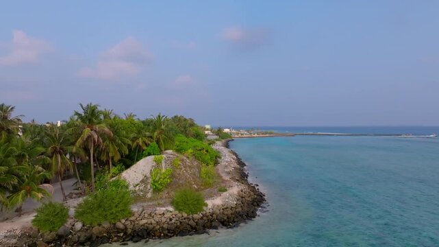 Aerial drone footage flying along the southern coast of Vashafaru Island where shoreline protection barriers line the beach, continuing toward the local harbor in northern Maldives.