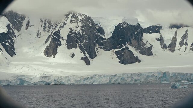 Antarctic glacial coastline with jagged snow-covered rocky mountains viewed through round ship porthole illuminator window