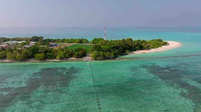 Aerial drone footage flying parallel to the southern coastline of Vashafaru Island featuring the local football field, harbor area and turquoise tropical waters in northern Maldives.