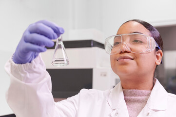Fototapeta premium Latina female researcher smiling as she observes a biochemical sample tube in the biotech lab wearing white coat and safety glasses.