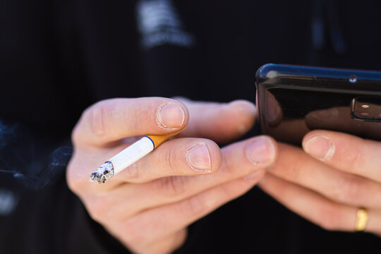 Close-up of hands holding a smartphone and a cigarette, with smoke rising, against a blurred background, showcasing a casual outdoor setting