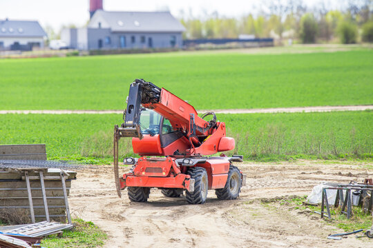 Red telescopic handler vehicle positioned on a farm with green fields and buildings in the background under a clear sky during daytime