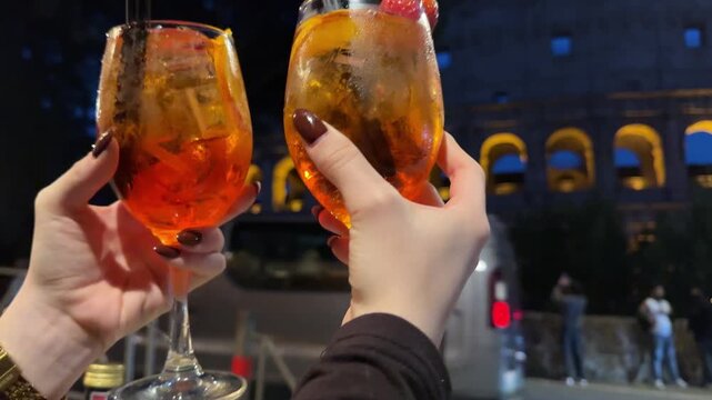 Close-up of female hands holding two Aperol cocktails against the illuminated Colosseum in Rome at night. A stylish travel and nightlife scene capturing atmosphere, drinks, and iconic landmark.