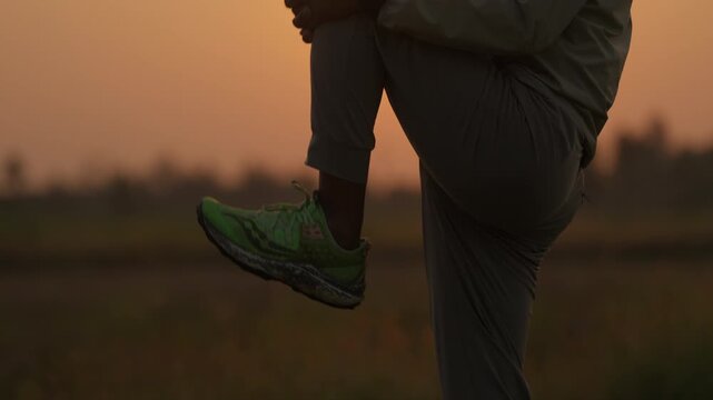 Golden light glows behind a person's hands. They stretch, moving slowly in open field. Sunset colors warm the horizon softly. Silhouette stands tall, back to camera