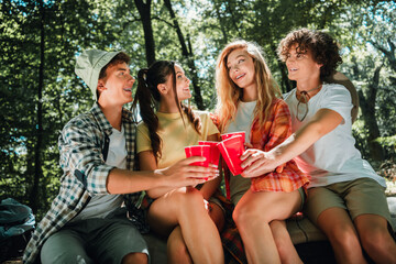 Obraz premium Four teenage friends camping in the woods sharing red cups, laughing and sitting together on a blanket during a sunny summer outdoor gathering