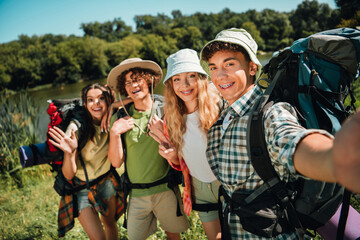 Teen friends on a summer backpacking trip take a happy selfie by a lake, smiling teenagers wearing...