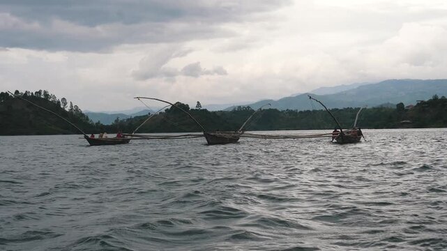 Three traditional wooden boats with fishermen casting a large net for fishing on the wavy water of lake kivu, with lush green hills and a cloudy sky in the background of rwanda