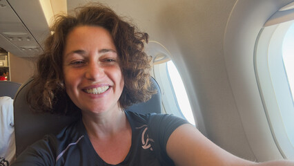 Naklejka premium Happy passenger young woman is flying in plane. Smiling girl sitting in airplane near window going on trip vacation travel. Traveling female inside plane enjoying flight. Traveling girl