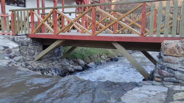 Small wooden bridge crossing a fast-flowing stream, surrounded by stones and rural architecture, creating a peaceful countryside scene.