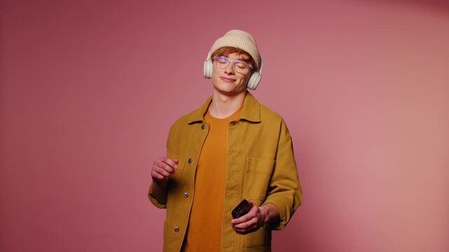Young man with glasses and headphones picks a song on his phone and dances against a pink background. He smiles and moves to the beat.