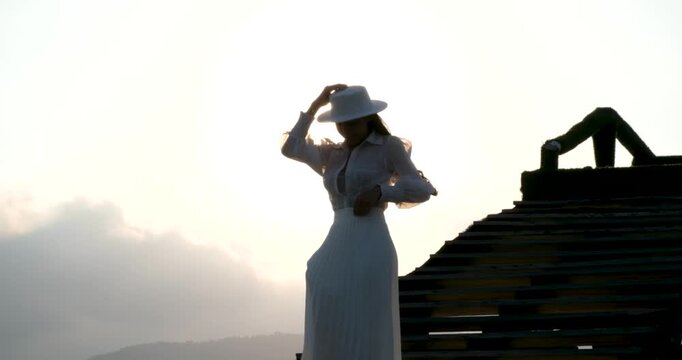 An Asian woman with long hair wearing a white blouse, white skirt, and hat walks along a wooden bridge at sunset. Warm golden sunlight illuminates her as she moves beside the ocean in a peaceful.
