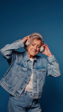 Happy senior woman in denim jacket and jeans enjoys music in headphones on blue background. Elderly female feeling joy and freedom.