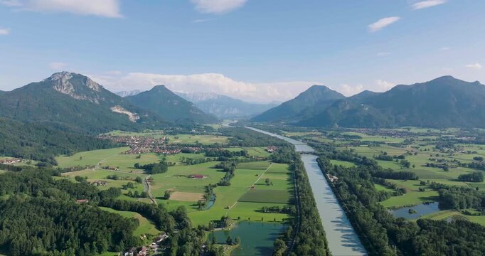 Aerial view of the tranquil Inn river flowing through a lush green valley, bordered by majestic mountains and a quaint village under a clear blue sky, Neubeuern, Bavaria, Germany.