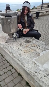 Samovar woman tea young woman sits by a traditional samovar at a seaside viewpoint drinking hot tea while wearing a warm ushanka hat
