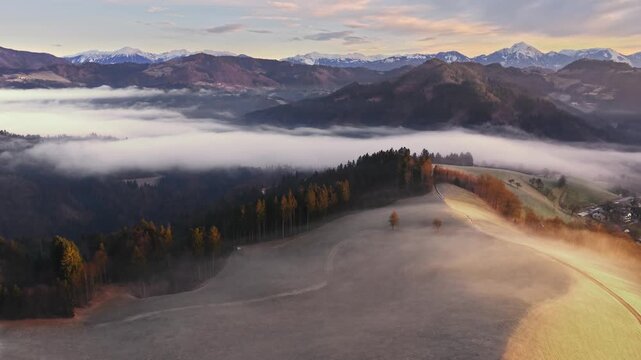 Aerial view of soft morning light paints rolling hills and a distant snow-capped mountain range through the fog, Admont, Styria, Austria.