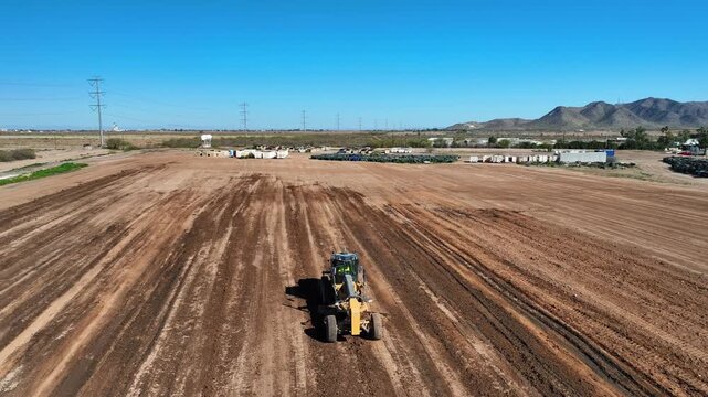 Aerial view of a vibrant yellow grader carving precise lines into the rich, reddish-brown earth, under a vast blue sky, Casa Grande, Arizona, United States.