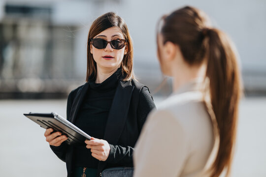 A businesswoman wearing sunglasses holds a tablet while speaking to a colleague. Two professionals converse outdoors in a brief business meeting scene.