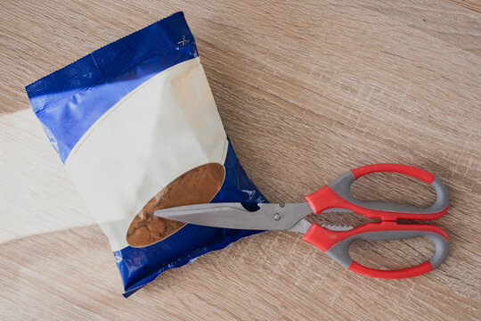 Package of cocoa powder and scissors on wooden kitchen table. Closed food package and kitchen scissors on wooden surface with natural light