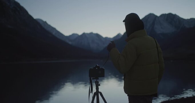 Photographer in a yellow jacket setting up a camera on a tripod by a calm mountain lake at dusk. Snow covered peaks, dark alpine valley, and soft reflections on the water create a moody outdoor scene