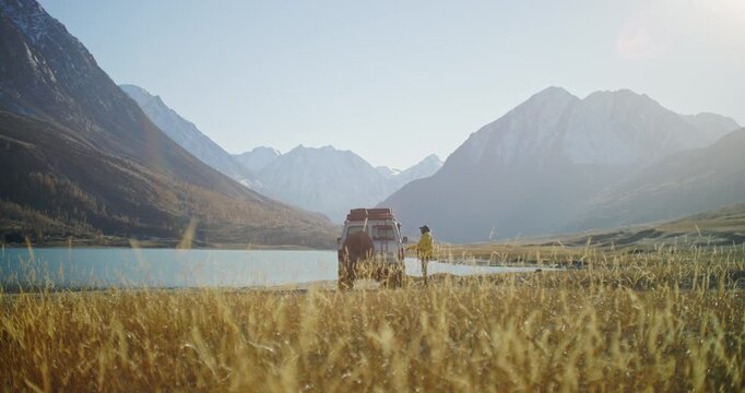 Traveler in a yellow jacket getting into an off road SUV parked by a mountain lake in a remote alpine valley. Golden grass, snowy peaks, and bright natural light create a scenic outdoor travel scene