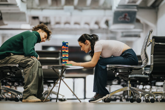 Two students focus on stacking a colorful block tower while seated across a low table. The casual scene shows concentration and playful teamwork in a modern lounge setting.