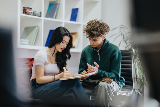 Students sit together reviewing notes and writing in a notebook. Two young people study and collaborate on schoolwork in a casual educational setting.