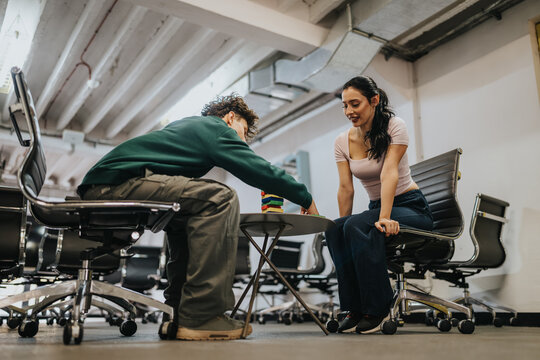 Two students play a stacking block game while seated in a modern office lounge. They sit on swivel chairs around a low table, concentrating on removing blocks without toppling the tower.
