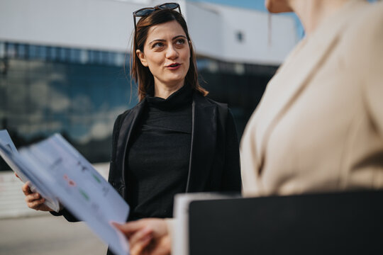Two businesswomen stand outside holding documents while discussing a project. They converse in a professional outdoor setting in front of a modern office building.